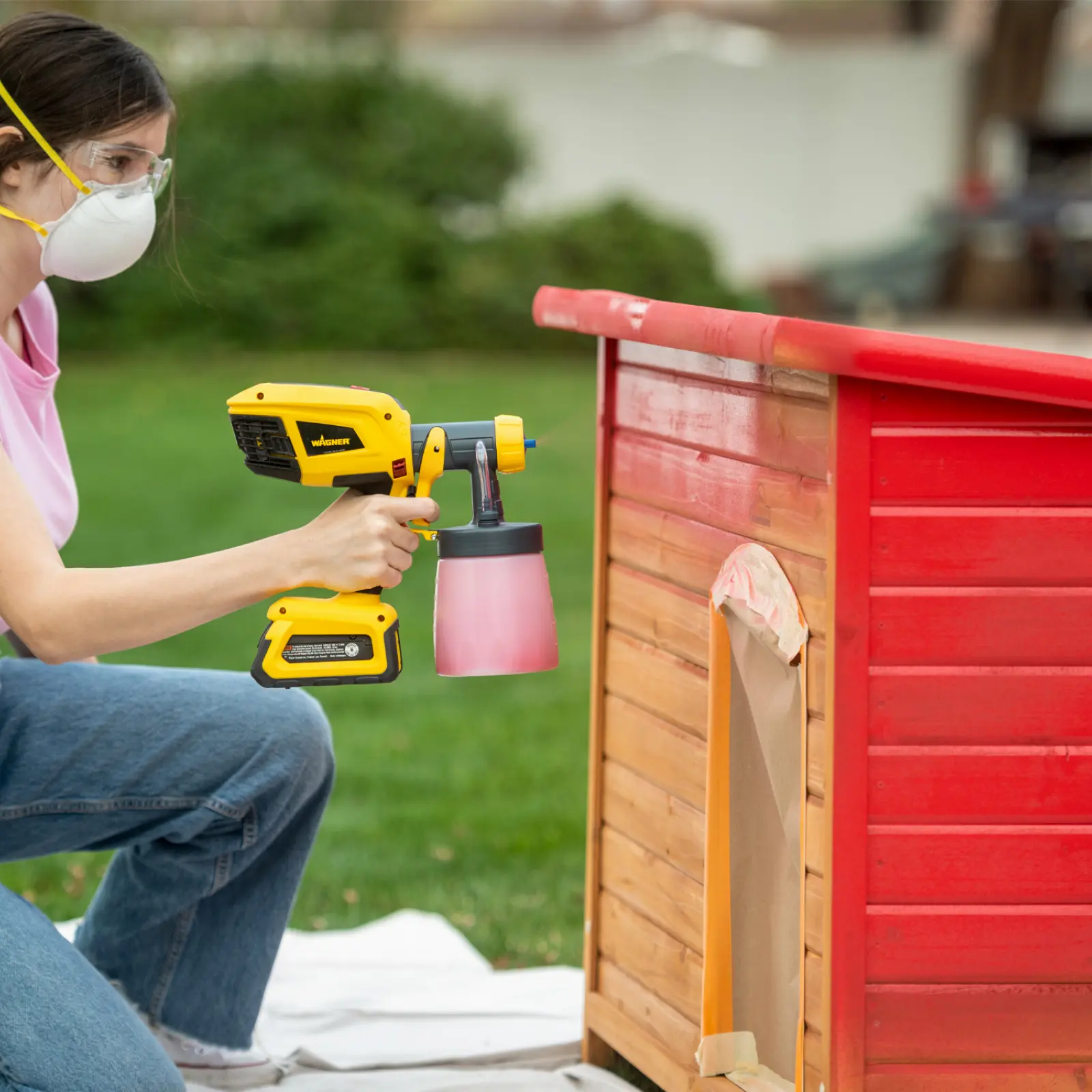 A woman using a cordless paint sprayer on a doghouse.