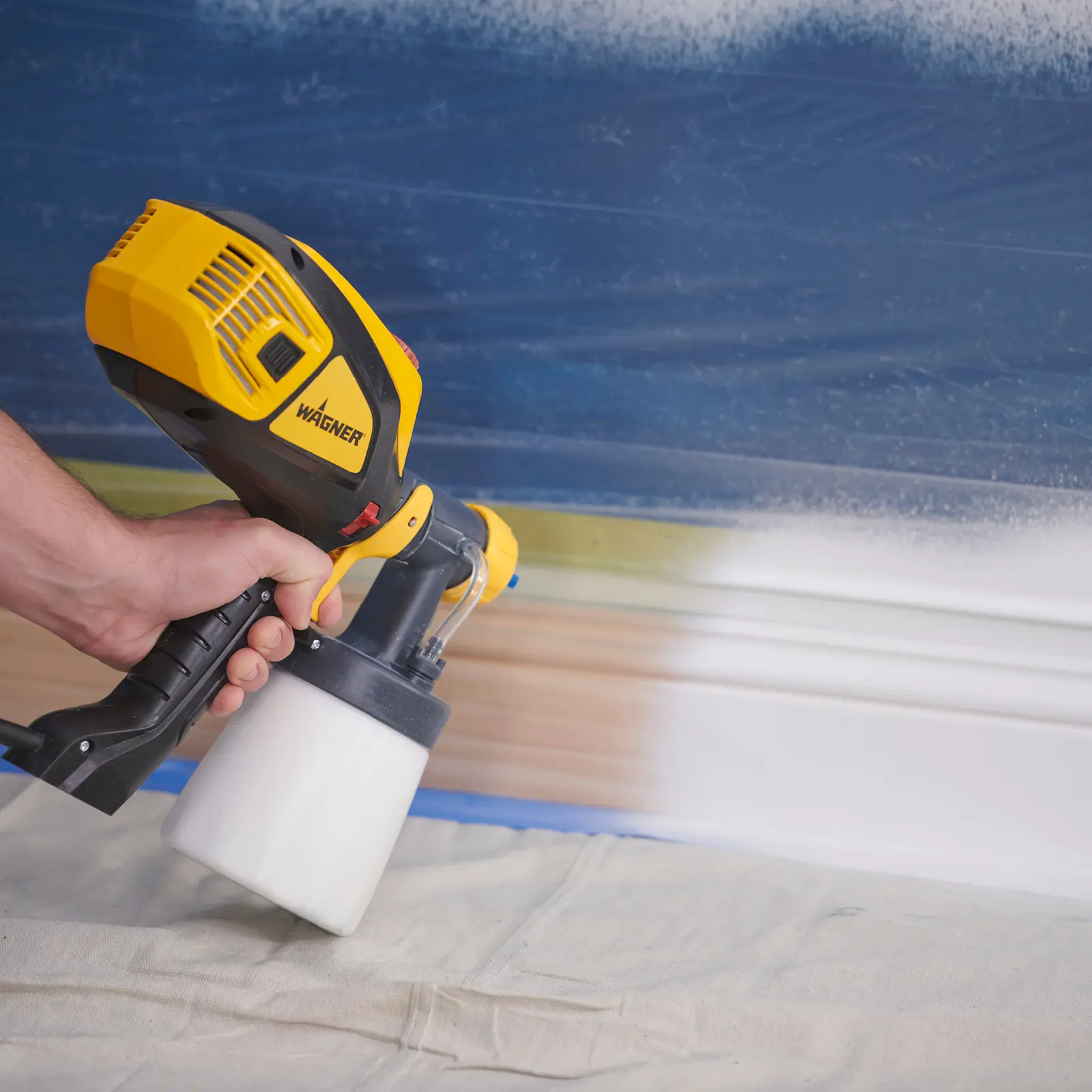Hand using a yellow Wagner paint sprayer on baseboards.