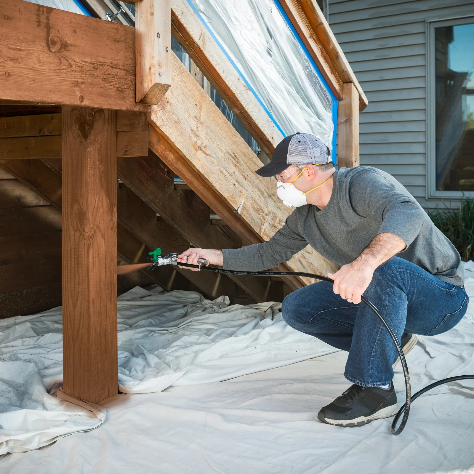 Man using a paint sprayer to stain a wooden deck.