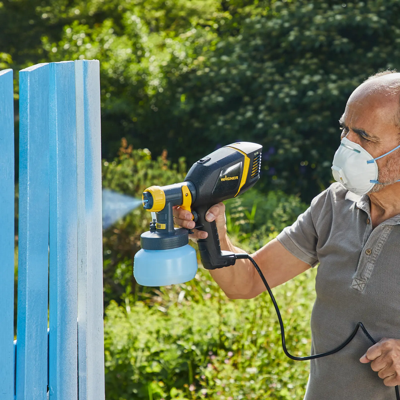 Man using a power paint sprayer to paint a fence.