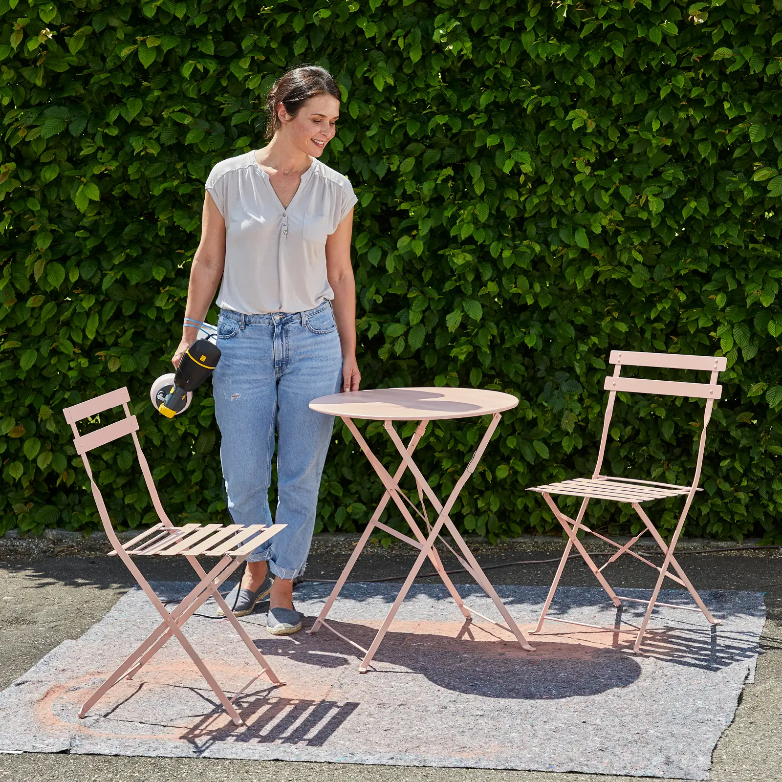 Woman using paint sprayer on pink patio furniture set.