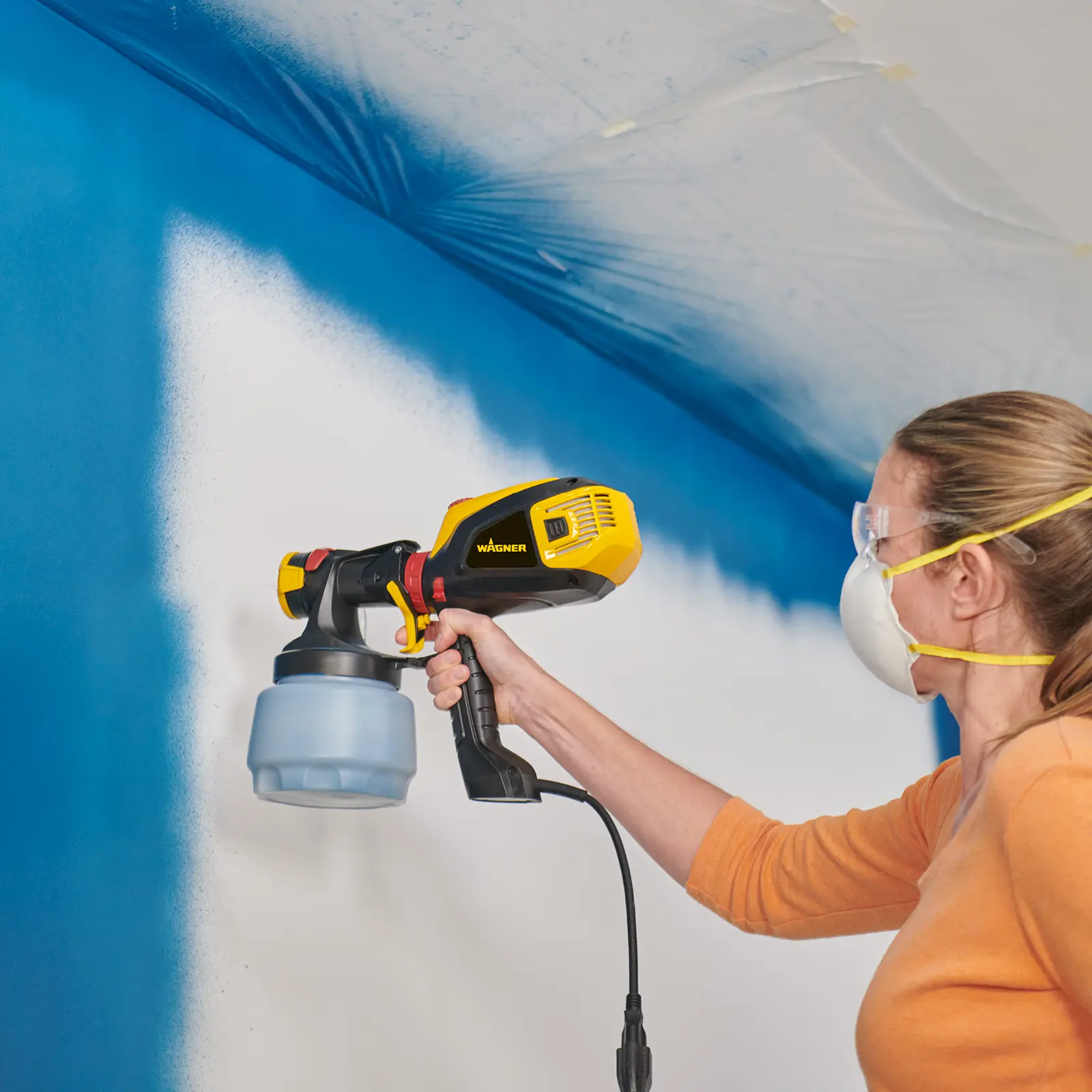 Woman using a yellow Wagner paint sprayer on a wall.