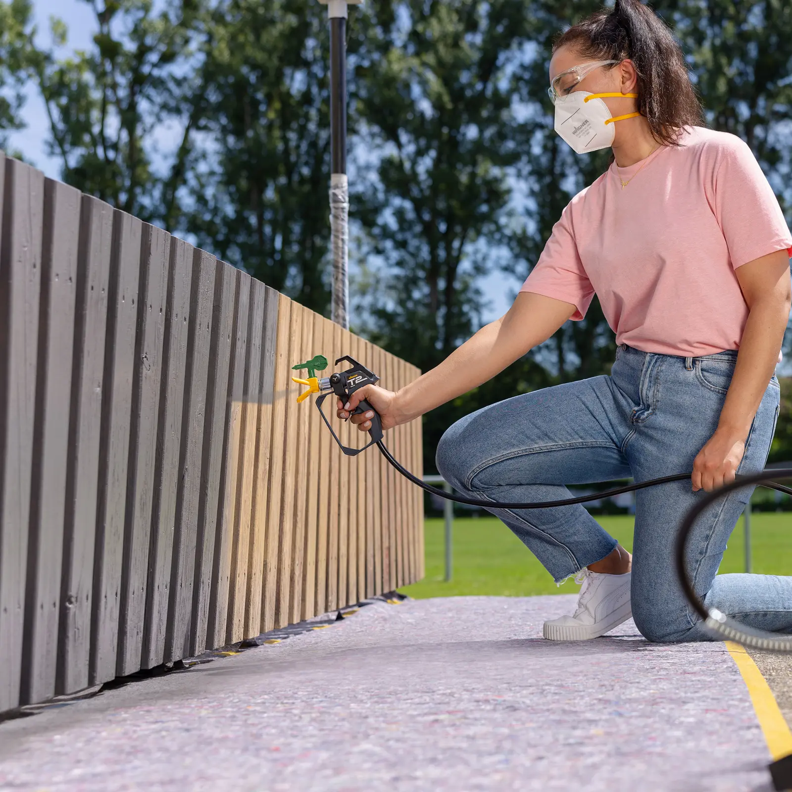Woman spray painting wooden fence outdoors with a paint sprayer.