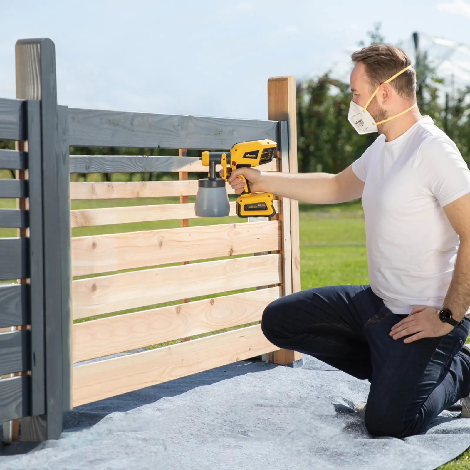 Man using a spray gun to paint a wooden fence gray.